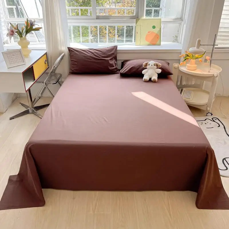 A neatly made bed with a mauve flat sheet, pillows, and a small stuffed animal on a light wood floor near a window.