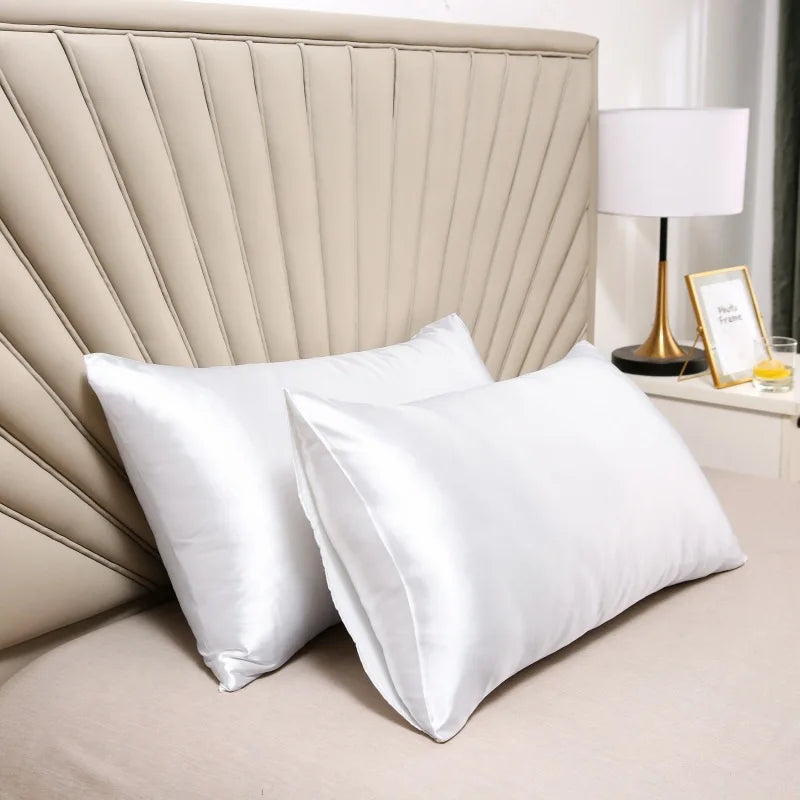 White satin silk pillow covers resting on a bed with a padded headboard and a lamp.