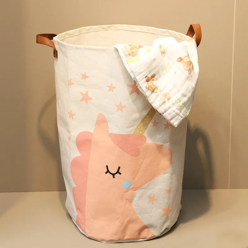 A light pink and white folding cartoon printed storage basket with a unicorn design and a fabric cloth attached.
