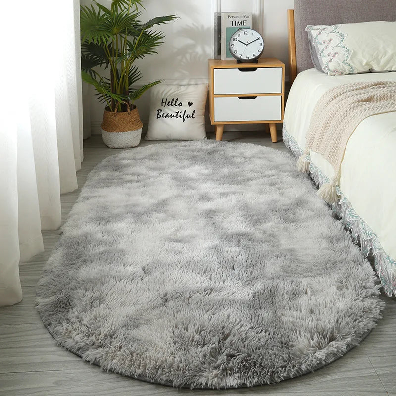 A fluffy gray plush floor mat placed in a bright room next to a wooden nightstand and a potted plant.