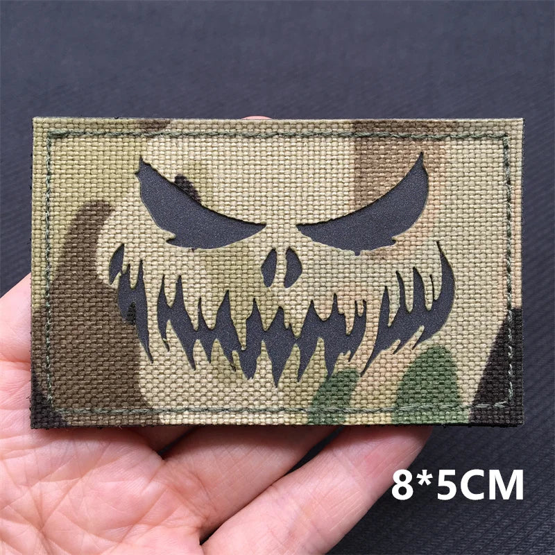 A rectangular tactical patch featuring a black skull design on a camouflage background, shown being held by a hand.