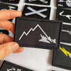 A hand holds a black embroidered patch featuring a white mountain line and a skull design.