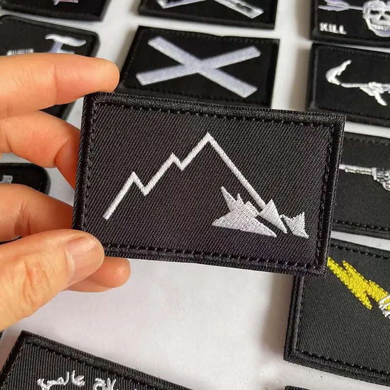 A hand holds a black embroidered patch featuring a white mountain line and a skull design.
