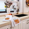 A pumpkin print kitchen towel draped over a sink counter next to various kitchen tools and seasonal decor.