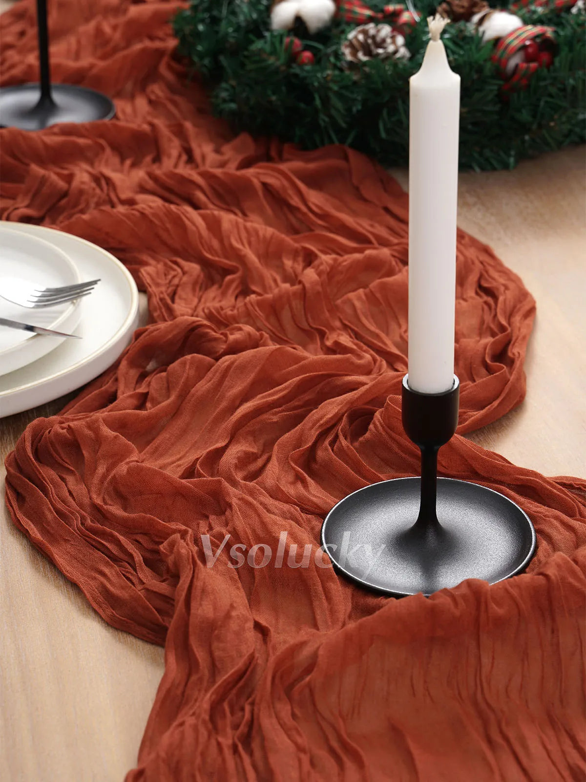 A terracotta orange crinkled cheesecloth table runner draped over a wooden surface with a white candle holder.