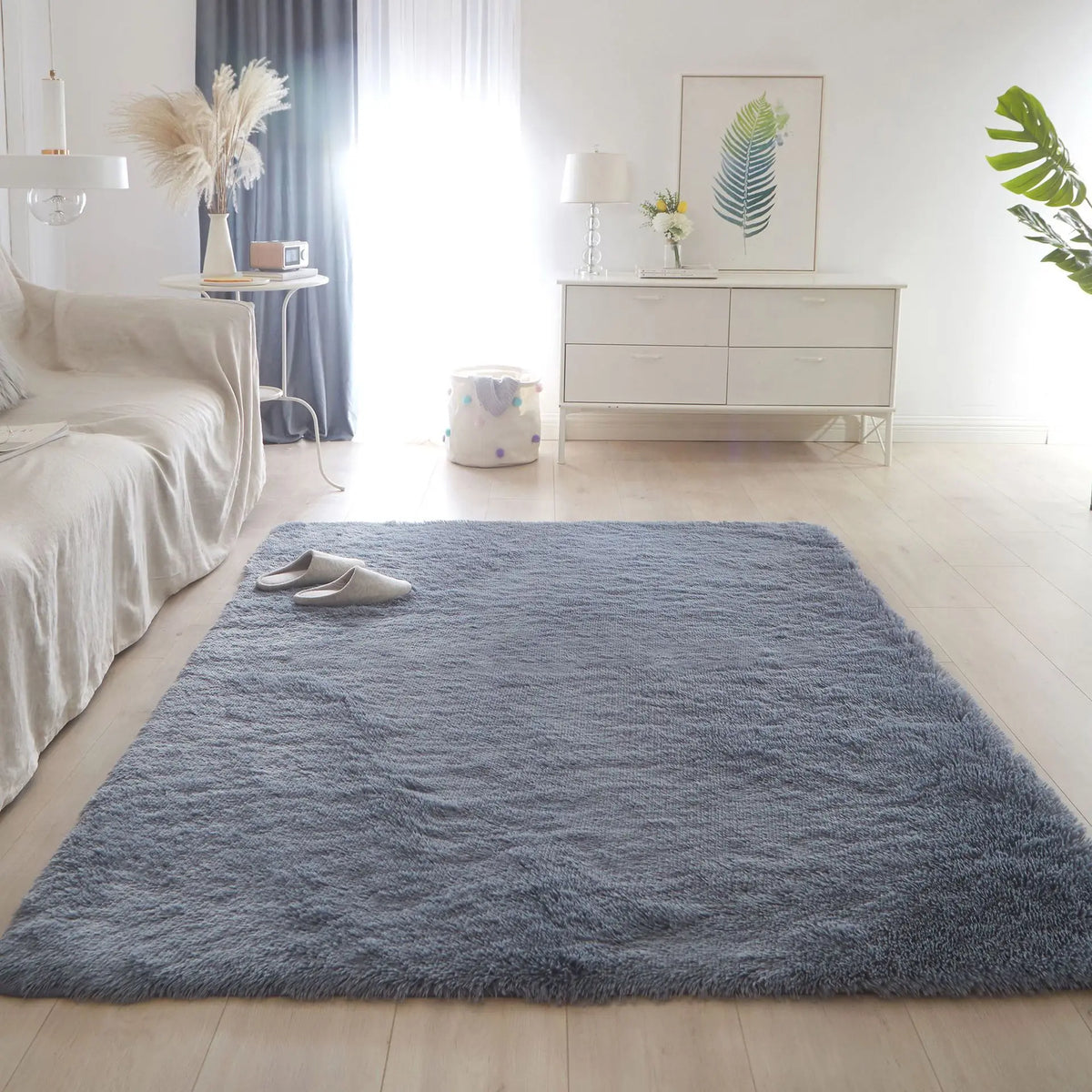 A fluffy blue gray shag rug placed on a light wood floor in a bright living room with white furniture and natural light.