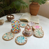 A set of five woven shell coasters with floral patterns arranged on a white table next to a potted plant and a candle.