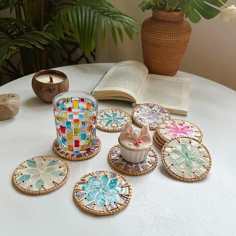 A set of five woven shell coasters with floral patterns arranged on a white table next to a potted plant and candle.
