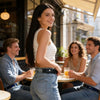 A young woman wearing a white top and jeans smiles while sitting at an outdoor cafe table.