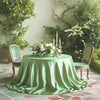A round table draped with a flowing sage green satin overlay cloth surrounded by greenery and white flowers.