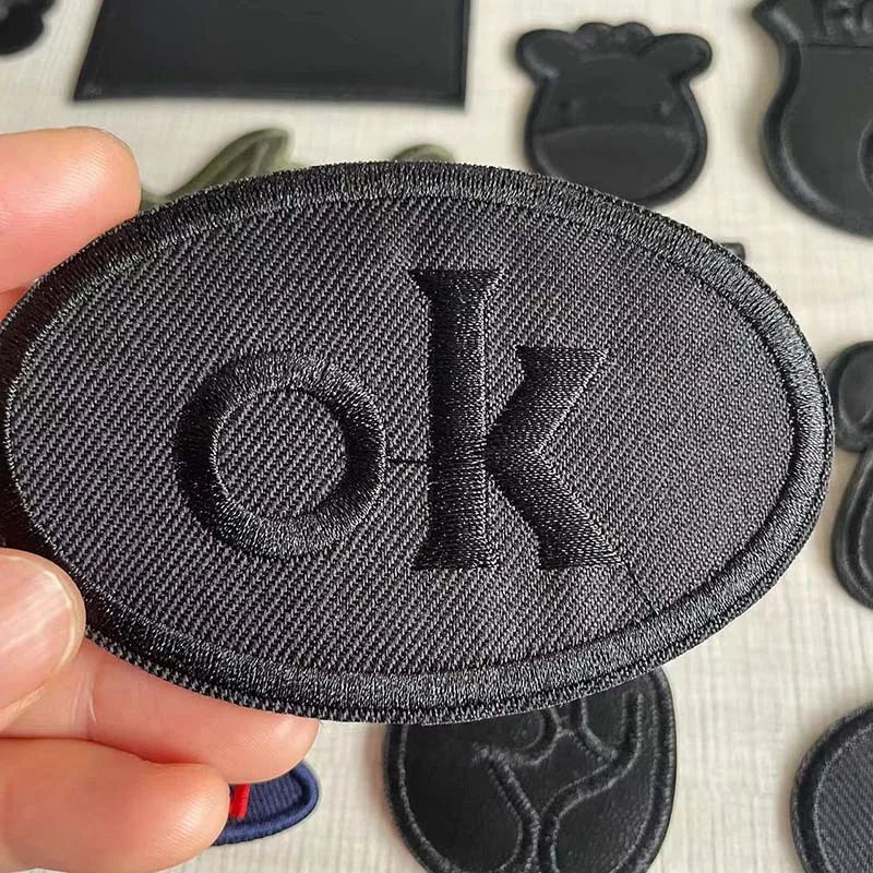 A hand holds a black embroidered leather patch with the letter OK on it against a white background.