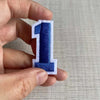 A hand holds a customizable number patch shaped like the numeral one, featuring blue and white embroidery.