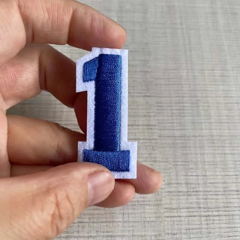 A hand holds a customizable number patch shaped like the numeral one, featuring blue and white embroidery.