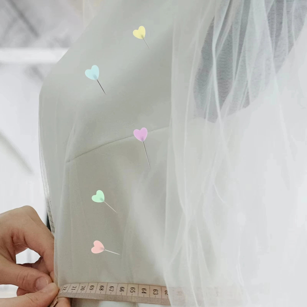 A close-up of a sheer fabric with heart-shaped sewing pins scattered across it, held by a hand.