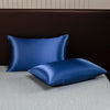 A pair of solid blue satin pillows resting on a light gray surface against a dark brown headboard.
