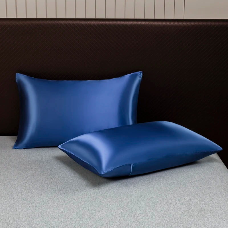 A pair of solid blue satin pillows resting on a light gray surface against a dark brown headboard.