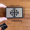 A black embroidered patch featuring a cross design on a light gray background, shown next to a hand and a hook.