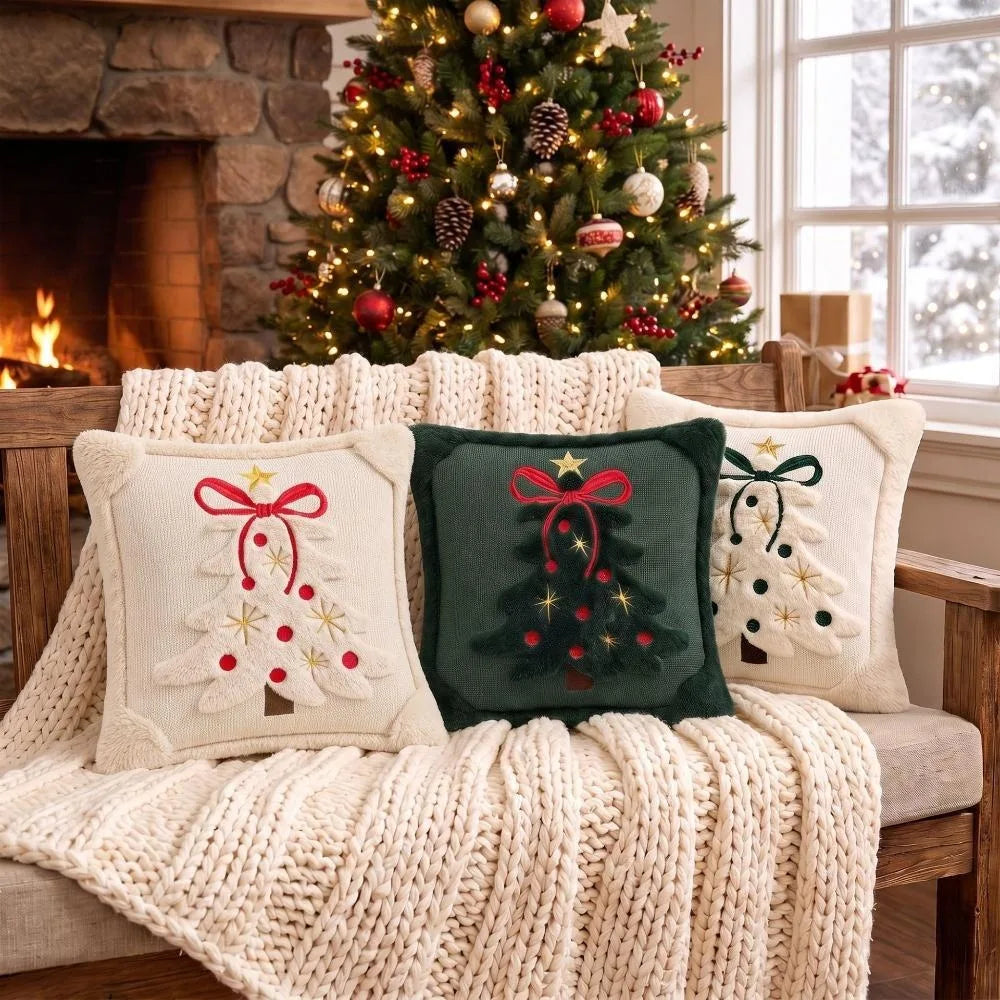 Three embroidered Christmas tree velvet pillow covers resting on a knitted blanket next to a decorated Christmas tree.