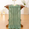 A sheer sage green cheesecloth table runner draped over a wooden table decorated with flowers.