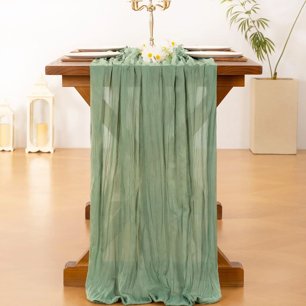 A sheer sage green cheesecloth table runner draped over a wooden table decorated with flowers.