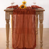A terracotta colored cheesecloth table runner draped over a wooden table with sunflowers and wine glasses.
