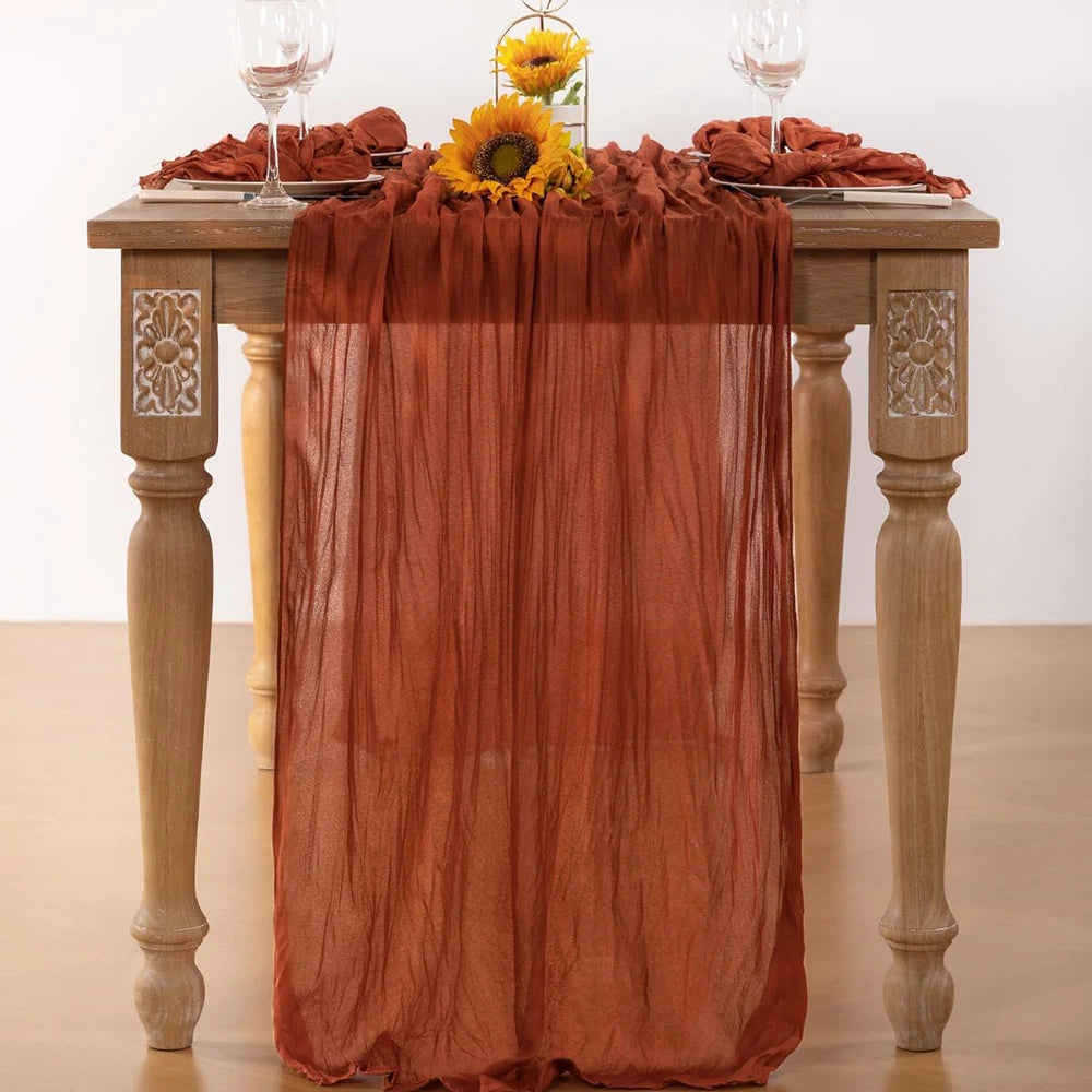 A terracotta colored cheesecloth table runner draped over a wooden table with sunflowers and wine glasses.