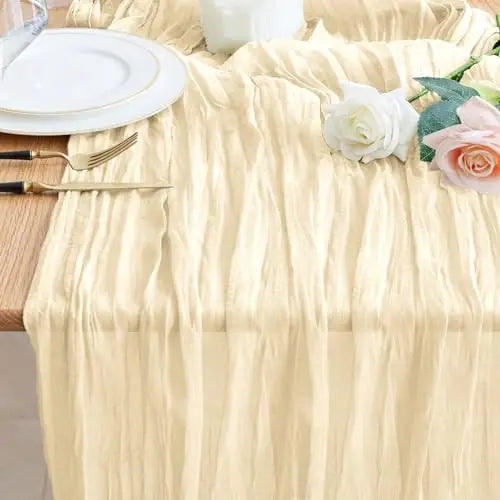 A beige cheesecloth table runner draped over a white tablecloth with white plates and floral decorations.