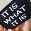 A close-up of a black embroidered patch with white text that reads IT IS WHAT IT IS being held by a hand.
