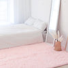 A pink shag rug is placed on a light wooden floor next to a white bed and a tall mirror.