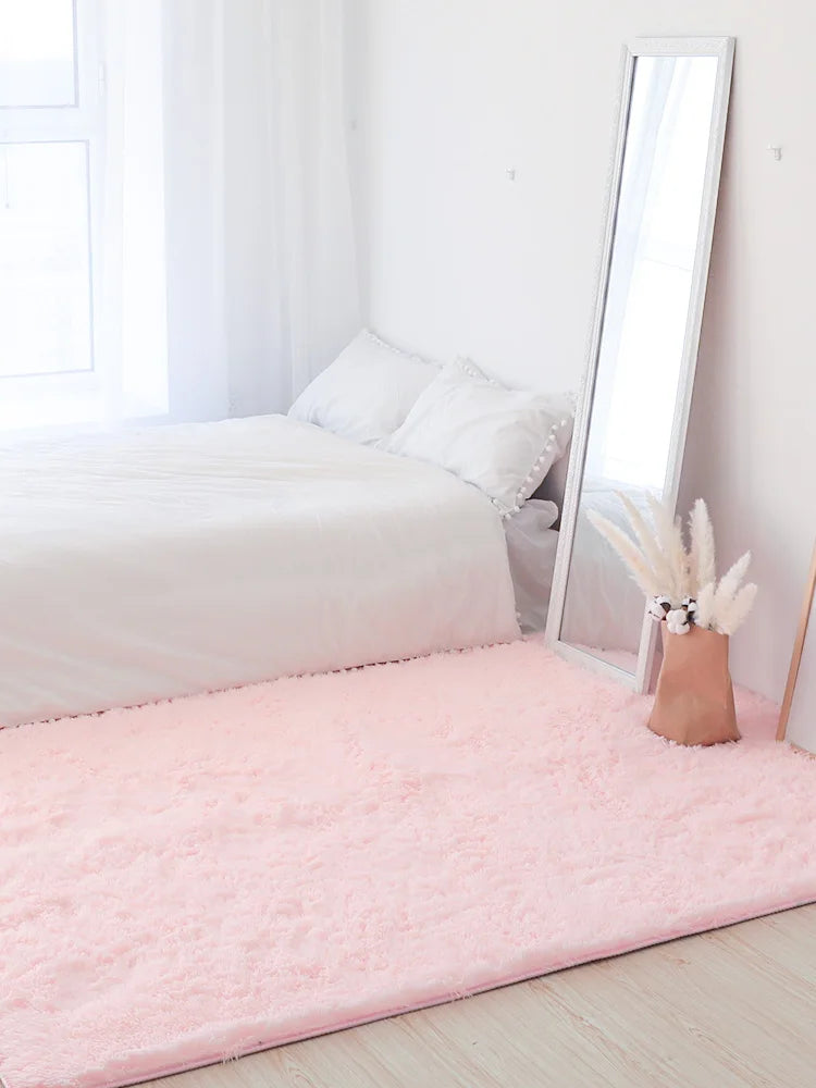 A pink shag rug is placed on a light wooden floor next to a white bed and a tall mirror.