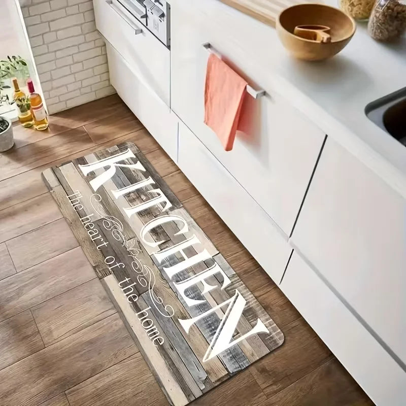 A wooden floor mat with carved text reading Kitchen the heart of the home placed on a light wood floor next to white kitchen 