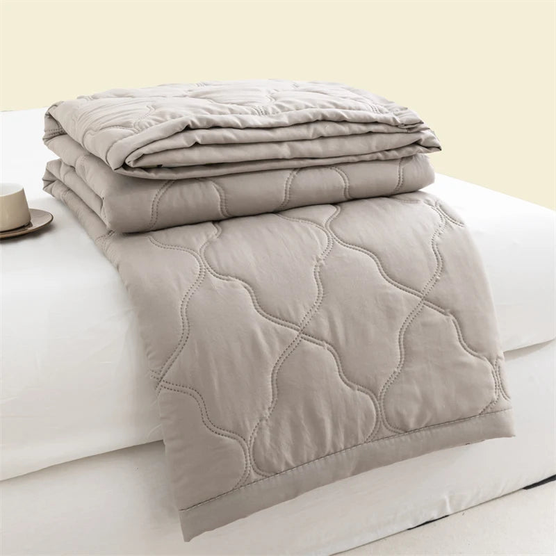 A stack of light gray quilted duvet covers and a folded duvet on a white bed.
