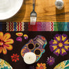 A dark table runner featuring a colorful skull and floral pattern, with a white candle resting on it.