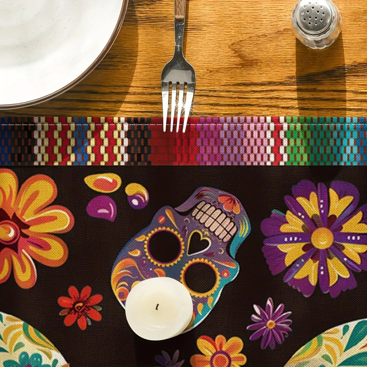 A dark table runner featuring a colorful skull and floral pattern, with a white candle resting on it.
