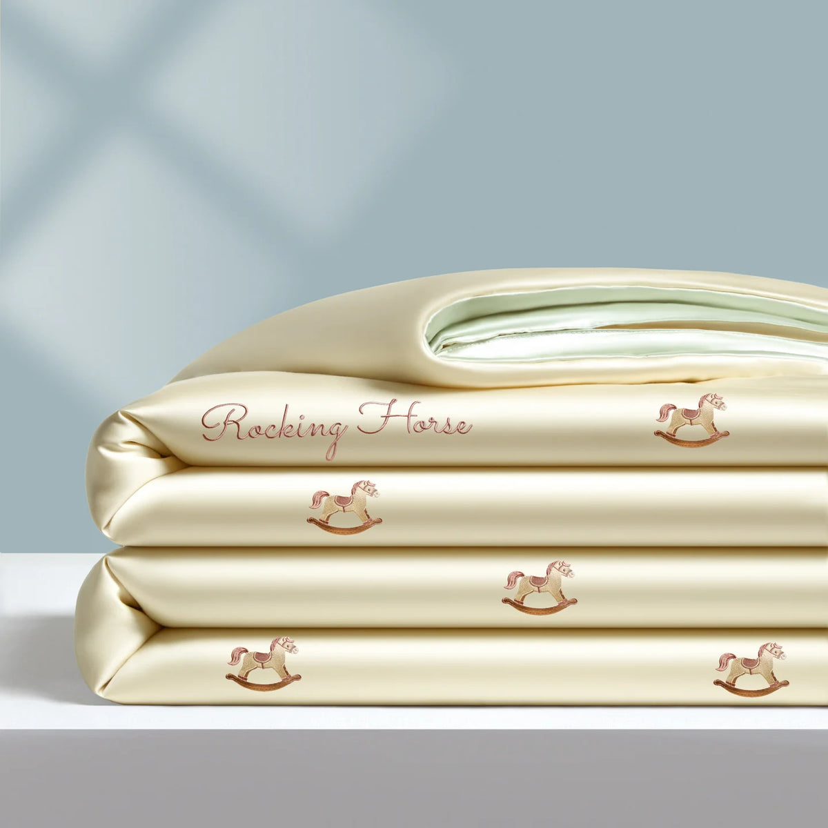 A stack of cream-colored cooling summer quilts embroidered with small pink rocking horses.