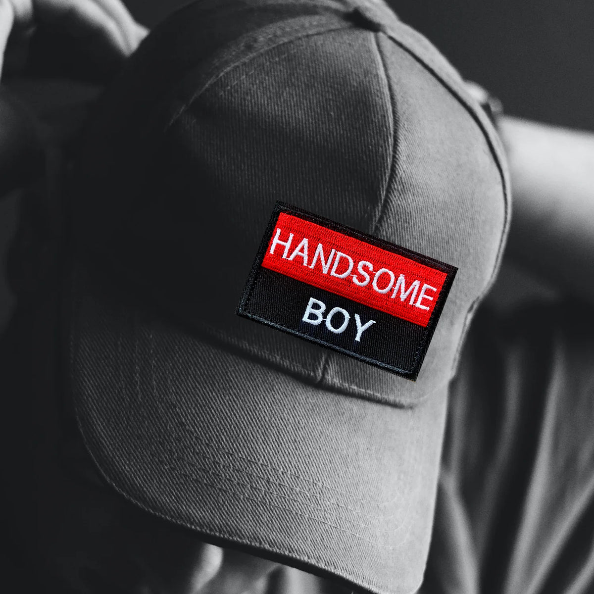 Close-up of a gray hat featuring a red embroidered patch with the text 'HANDSOME BOY'
