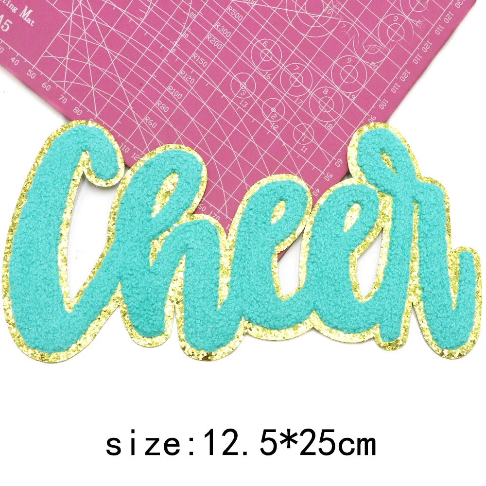 A turquoise glitter script letter cheer patch on a pink grid background.