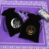 Two black velvet tarot cards storage pouches with gold and silver designs resting on a purple patterned cloth.