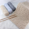 A beige knitted rug is displayed next to three skeins of gray and light blue yarn and a pair of wooden chopsticks.