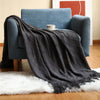A dark gray knitted tassel throw blanket draped over a blue armchair on a wooden floor.