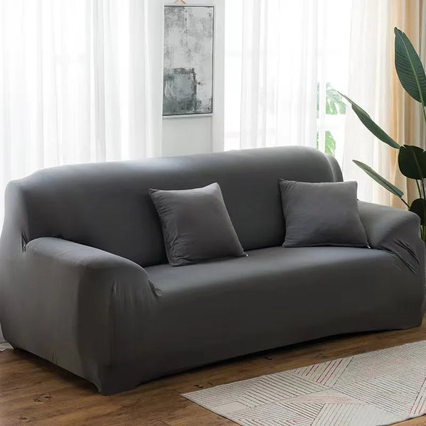 A modern gray stretch sofa cover placed in a bright living room with sheer white curtains and a wooden floor.