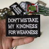 A hand holds a black iron-on slogan patch with white text that reads Don't mistake my kindness for weakness.