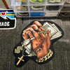 A hand is holding a gold chain and a cross over stacks of money on a black background.