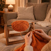 Hands holding orange yarn on a wooden rotating storage rack next to a sofa.