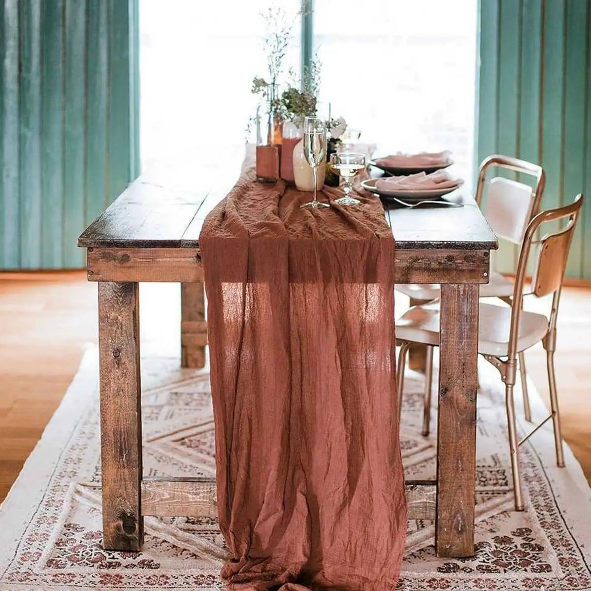 A rustic wooden table set with a sheer terracotta gauze table runner and elegant place settings.