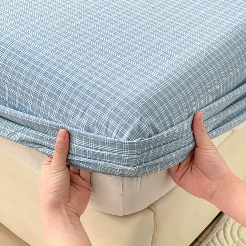 A hand is holding up a light blue plaid fitted sheet on a white bed frame.