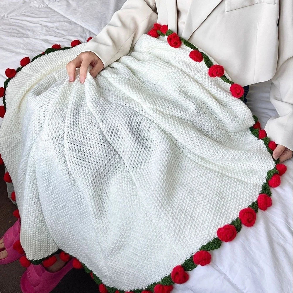 A white knitted blanket with a green border and red floral accents is being held on a white bed.