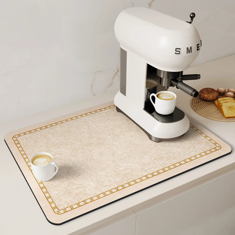 A white electric coffee maker is placed on a beige drain mat with gold trim, next to a cup of coffee.