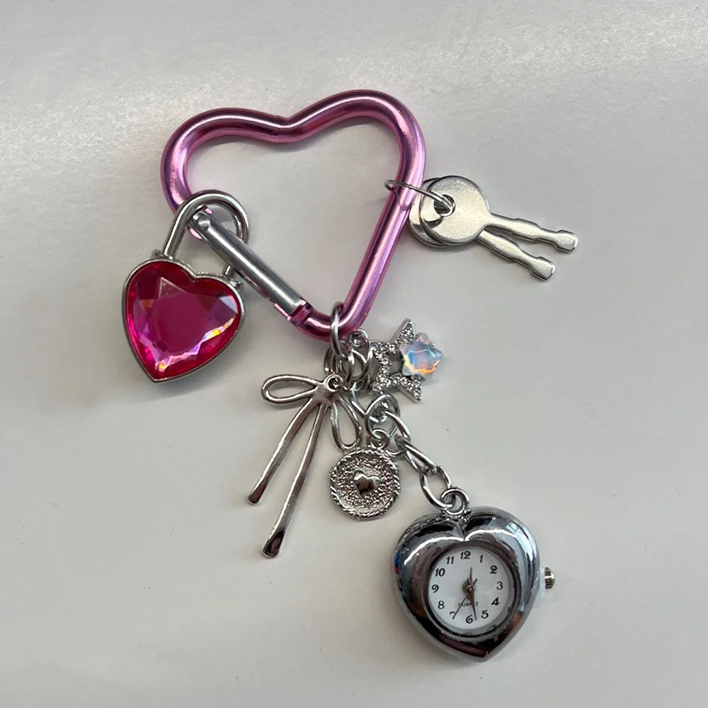 A pink heart-shaped keychain featuring a pink heart charm, a silver tag, and a small silver pocket watch.
