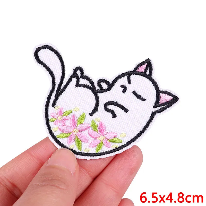 A hand holds a pink and white embroidered patch of a sleeping cat with floral details.